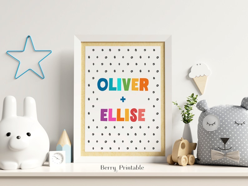 Op de afbeelding: Een ingelijste print met de namen "OLIVER + ELLISE" in kleurrijke letters op een witte achtergrond met zwarte stippen. De print staat op een witte plank met een ster, een konijn en een beer, ideaal voor een kinderkamer.