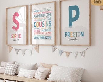 May include: Three framed art prints with wooden frames hang on a white wall. The prints feature the letters "S" and "P" with names and the word "Cousins" with a list of activities. A garland of small flags hangs below the prints.
