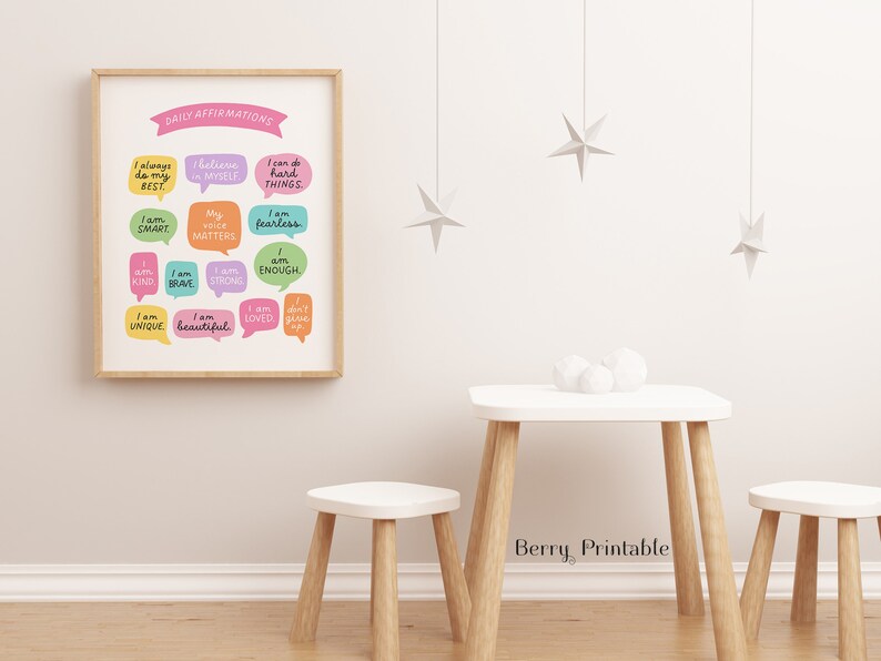 May include: A printable poster with a light wood frame featuring a colourful design of speech bubbles with positive affirmations. The poster is hanging on a white wall with a small white table and two white stools in front of it.