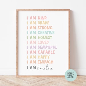 May include: A framed print with a pastel rainbow color scheme. The print features the text "I am" followed by a list of positive affirmations, including "kind", "brave", "strong", "creative", "honest", "loved", "beautiful", "capable", "happy", and "enough". The print is personalized with the name "Emilia".
