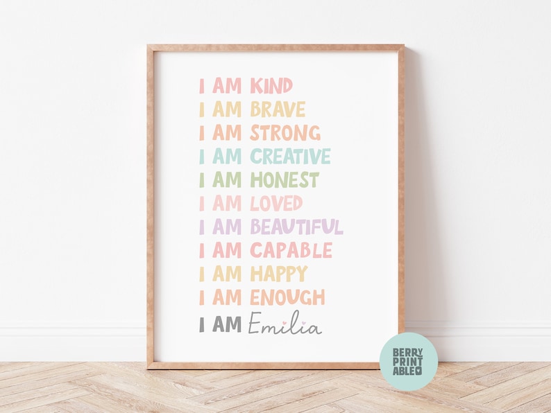 May include: A framed print with a pastel rainbow color scheme. The print features the text "I am" followed by a list of positive affirmations, including "kind", "brave", "strong", "creative", "honest", "loved", "beautiful", "capable", "happy", and "enough". The print is personalized with the name "Emilia".