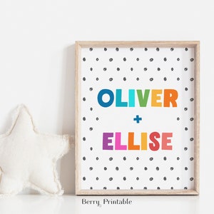 Op de afbeelding: Een ingelijste print met de namen "OLIVER + ELLISE" in kleurrijke blokletters. De achtergrond is wit met zwarte stippen. Links van de lijst ligt een wit stervormig kussen.
