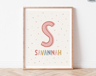 May include: A framed print featuring the name "Savannah" in colorful letters below a large pink letter "S". The background is off-white with scattered pastel-colored dots. The print is in a light brown wooden frame.