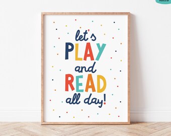 May include: A rectangular wall art print in a light wood frame featuring the text let's PLAY and READ all day! in dark blue, yellow, red, and teal lettering. The white background is decorated with small, scattered multicolored polka dots.