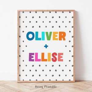 Op de afbeelding: Een ingelijste print met de namen "OLIVER" en "ELLISE" in kleurrijke, dikgedrukte letters. De namen staan tegen een witte achtergrond met een patroon van kleine, zwarte stippen. De lijst is lichtbruin.