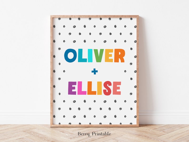 Op de afbeelding: Een ingelijste print met de namen "OLIVER" en "ELLISE" in kleurrijke, dikgedrukte letters. De namen staan tegen een witte achtergrond met een patroon van kleine, zwarte stippen. De lijst is lichtbruin.
