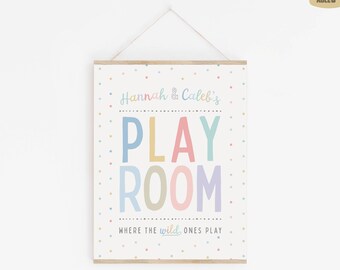 May include: A rectangular wall hanging sign with a white background featuring colorful polka dots. The text reads Hannah & Caleb's PLAY ROOM WHERE THE wild ONES PLAY in various pastel colors. The sign is suspended by a thin string from a wooden frame.