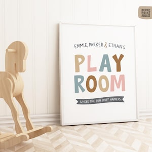 May include: A white framed print with the text "EMMIE, PARKER & ETHAN'S PLAY ROOM WHERE THE FUN STUFF HAPPENS" in colorful letters. The letters are in shades of brown, pink, and blue.