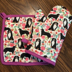 May include: A set of two pink and white oven mitts with a floral pattern and Bernese Mountain Dog illustrations. The mitts are quilted and have a hanging loop.