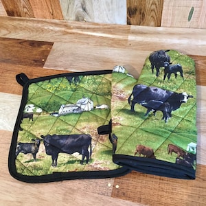 May include: A set of two black and green quilted kitchen pot holders with a farm scene featuring cows, a barn, and a green field. The pot holders have black trim and a hanging loop.