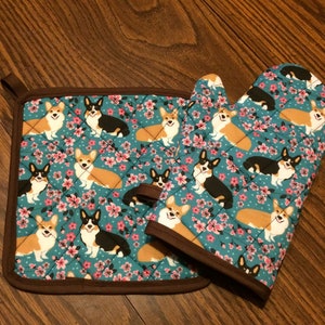 May include: A set of two teal blue oven mitts and a pot holder with a pattern of brown and black corgis on a teal blue background with pink flowers.