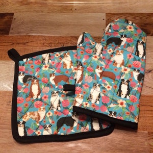 May include: A set of two blue and black oven mitts and a pot holder with a floral pattern and a repeated image of Australian Shepherd dogs.