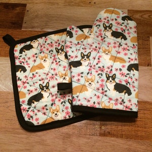 May include: A quilted oven mitt and pot holder set. The light blue fabric features a repeating pattern of corgi dogs and pink flowers. The set is trimmed with black binding.