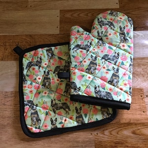 Puede incluir: Un juego de manopla y agarradera acolchadas. El juego presenta un fondo verde claro con un patrón repetido de perros ganaderos australianos y flores rosas. Los bordes están ribeteados con una encuadernación negra.