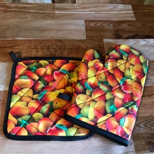 May include: A quilted pot holder and oven mitt set. The set features a vibrant pattern of peaches in shades of red, yellow, and green. The edges are trimmed with black binding. The pot holder has a black loop for hanging.