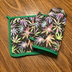 May include: A set of two green-trimmed pot holders with a black background and a colorful marijuana leaf pattern.
