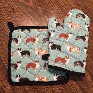 May include: A quilted oven mitt and pot holder set. The light blue fabric features a repeating pattern of Australian Shepherd dogs in various colours. The set is trimmed with black binding.