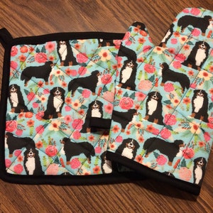 May include: A set of two quilted oven gloves featuring a floral pattern with Bernese Mountain Dog illustrations. The oven gloves are blue with pink flowers and black dogs.