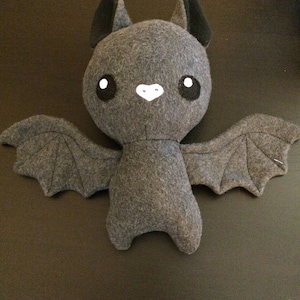 May include: A gray felt bat plush toy with black ears and wings. The bat has large, round eyes with white highlights and a small white nose. The wings are spread out, and the toy is on a dark surface.