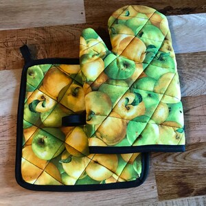 Puede incluir: Un juego de dos agarraderas de cocina acolchadas con estampado de manzanas verdes y amarillas. Las agarraderas tienen un ribete negro.