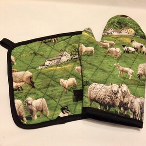 以下が含まれることがあります： 緑と白の農場をテーマにした、キルティングされた鍋つかみの2点セット。鍋つかみは黒い縁取りと、羊と農家のパターンが特徴です。