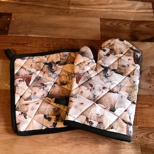 Op de afbeelding: Een set van twee gewatteerde ovenwanten met een zwarte rand. De ovenwanten hebben een witte achtergrond met een herhalend patroon van schapengezichten.