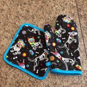 Peut inclure: Un ensemble de deux maniques de four noires avec une bordure turquoise. Les maniques présentent un motif coloré inspiré du Jour des Morts avec des squelettes de chiens portant des fleurs.