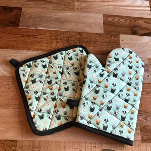 May include: A quilted oven mitt and pot holder set. The set features a light green background with a repeating pattern of French bulldog faces in black, white, and brown. The edges are trimmed with black binding.
