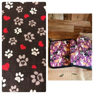 Puede incluir: Un juego de guantes para horno de color morado y negro y un posavasos con un patrón de corgis bebiendo vino. Los guantes y el posavasos tienen un borde negro.