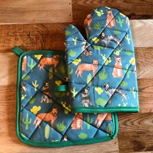 Puede incluir: Un juego de dos manoplas para horno azules y verdes con un patrón de pastores australianos y cactus. Las manoplas están acolchadas y tienen un ribete verde.