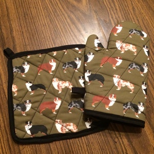 May include: A set of two green oven mitts with a black trim. The mitts are decorated with a pattern of Australian Shepherd dogs.