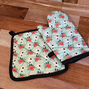 Puede incluir: Un juego de dos agarraderas de cocina con fondo azul claro y un patrón de caras de bull terrier blanco y rosas rosas. Las agarraderas tienen un ribete negro.