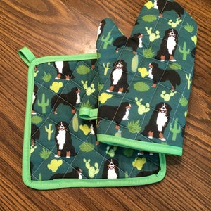 May include: A green quilted oven mitt and pot holder set with a pattern of Bernese Mountain Dogs and cacti. The set has a green trim.