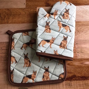 May include: A set of two kitchen pot holders featuring a pattern of German Shepherd dogs on a light blue background. The pot holders have brown trim.