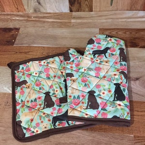 Könnte beinhalten: Ein Set aus zwei Topflappen mit hellblauem Hintergrund und einem Muster aus schwarzen und gelben Labrador Retrievern und rosa Blumen. Die Topflappen haben einen braunen Rand.