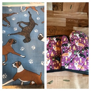 May include: Two oven mitts with a purple background and a pattern of corgis and wine glasses. The mitts are quilted and have a black trim.