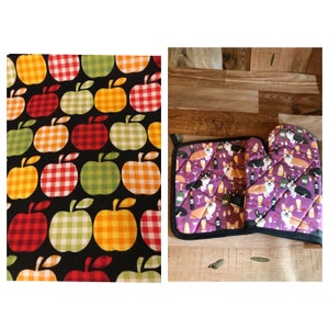 Puede incluir: Un juego de dos agarraderas de cocina. Una es de patrón a cuadros rojo y blanco con manzanas y la otra es una agarradera morada con un patrón de corgis y copas de vino.
