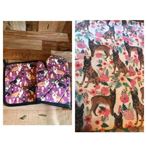 May include: A set of two purple oven mitts with a black trim. The mitts feature a pattern of corgis and wine glasses. The mitts are on a wooden surface.