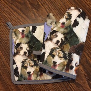 May include: A set of two quilted oven gloves and a pot holder featuring a colourful pattern of Australian Shepherd dogs. The gloves and pot holder have a grey trim.