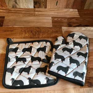 May include: A matching oven mitt and pot holder set. The items display a repeating pattern of black and brown dogs on a light grey background. Both are edged with black fabric. The pot holder has a hanging loop.