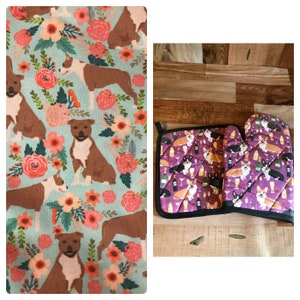 May include: A set of two kitchen pot holders featuring a purple background with a repeating pattern of corgis drinking wine. The pot holders are made of quilted fabric and have a black trim.