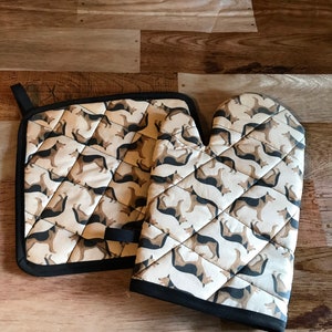 May include: A set of two brown and black patterned oven mitts. The mitts feature a repeating pattern of German Shepherd dogs. The mitts are quilted and have a black trim.