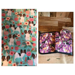 May include: A set of kitchen accessories featuring a teal blue tea towel with a repeating pattern of white and brown papillon dogs and pink flowers, and a purple pot holder and oven mitt with a repeating pattern of brown and white corgi dogs, wine glasses, and wine bottles.