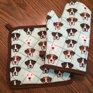 May include: A set of two blue and brown oven mitts with a pattern of boxer dog faces. The mitts are quilted and have a brown trim.