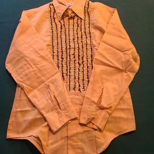 May include: A long-sleeved, button-down shirt in a light orange hue. The shirt features a ruffled design in black and gold down the front, with a classic collar and buttoned cuffs. The fabric appears to be a lightweight material.