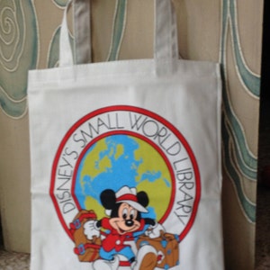 May include: White canvas tote bag with a red, white, and blue logo featuring Mickey Mouse holding luggage. The logo says "Disney's Small World Library".