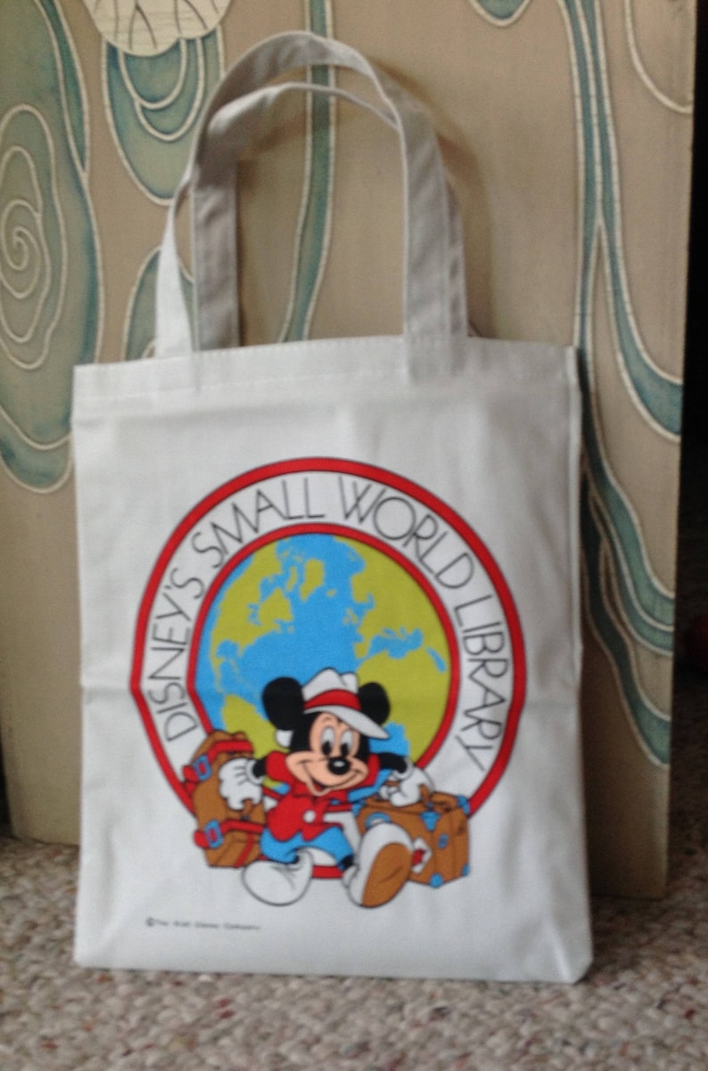 May include: White canvas tote bag with a red, white, and blue logo featuring Mickey Mouse holding luggage. The logo says "Disney's Small World Library".