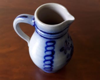 May include: A small ceramic pitcher with a light gray glaze and dark blue hand-painted accents. The vessel features a rounded body, a curved handle, and a pouring spout, decorated with vertical blue oval patterns and floral motifs.