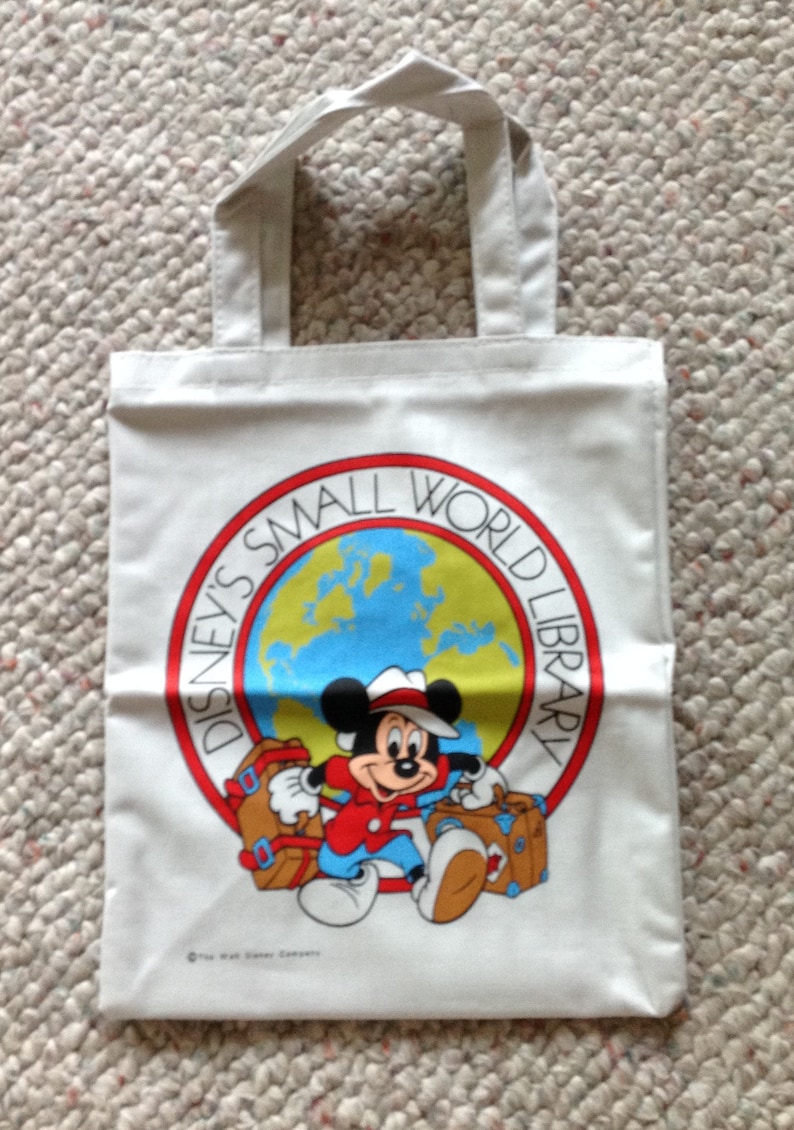 May include: White canvas tote bag with a red, yellow, and blue circle design featuring Mickey Mouse holding luggage. The circle says "Disney's Small World Library".