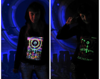 Peut inclure: Un t-shirt noir à manches longues avec un motif qui brille dans le noir. Le motif représente un cercle avec un cercle plus petit à l'intérieur, entouré d'un anneau arc-en-ciel. Le t-shirt est présenté en deux images, l'une sous une lumière noire UV et l'autre dans le noir. Le texte "triple view art" est visible dans le coin supérieur droit de l'image.